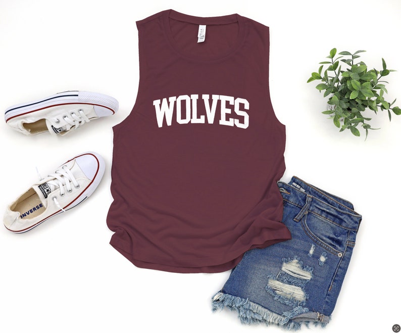 May include: A maroon sleeveless shirt with the word "WOLVES" in white block letters. The shirt is paired with denim shorts and white canvas sneakers. A small green plant is also in the image.