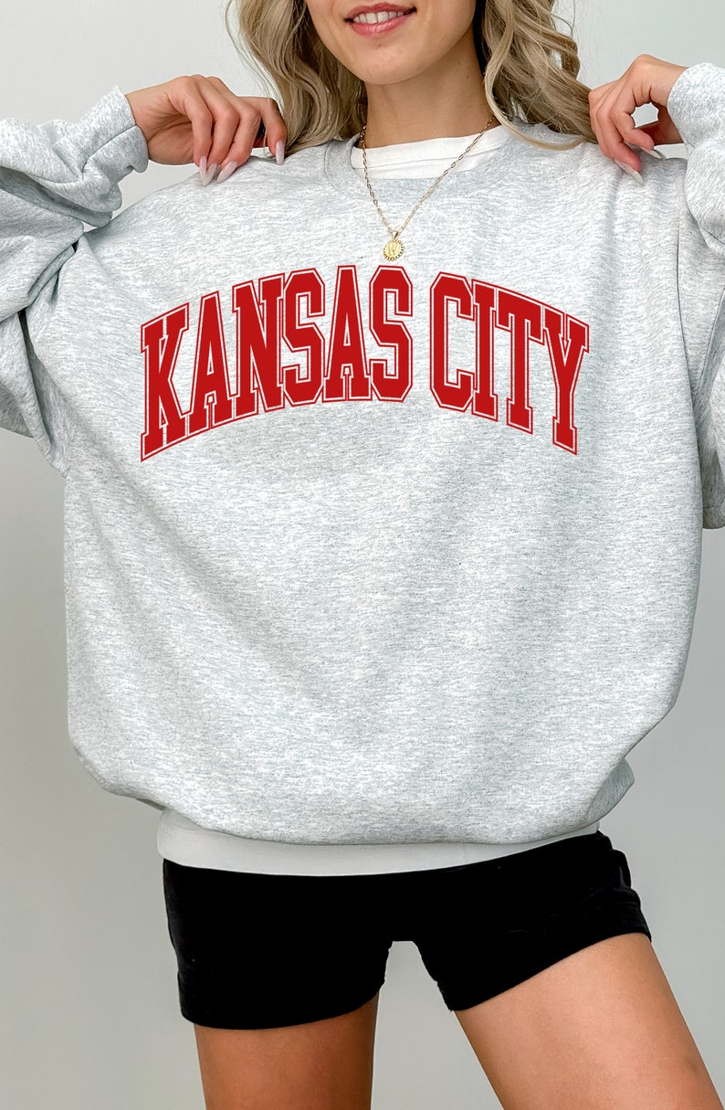 Op de afbeelding: Een grijs sweatshirt met de tekst "KANSAS CITY" in rode letters.