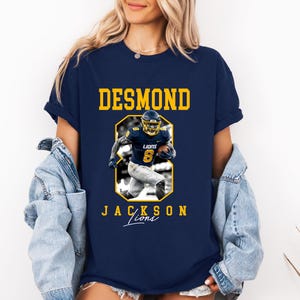 May include: A navy blue short-sleeve t-shirt featuring a graphic of a football player in a blue and yellow uniform. The text DESMOND is printed in yellow above the player, and JACKSON LIONS is printed below in yellow with the word Lions in script.