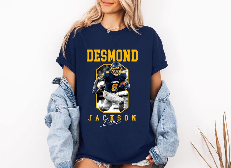 May include: A navy blue short-sleeve t-shirt featuring a graphic of a football player in a blue and yellow uniform. The text DESMOND is printed in yellow above the player, and JACKSON LIONS is printed below in yellow with the word Lions in script.