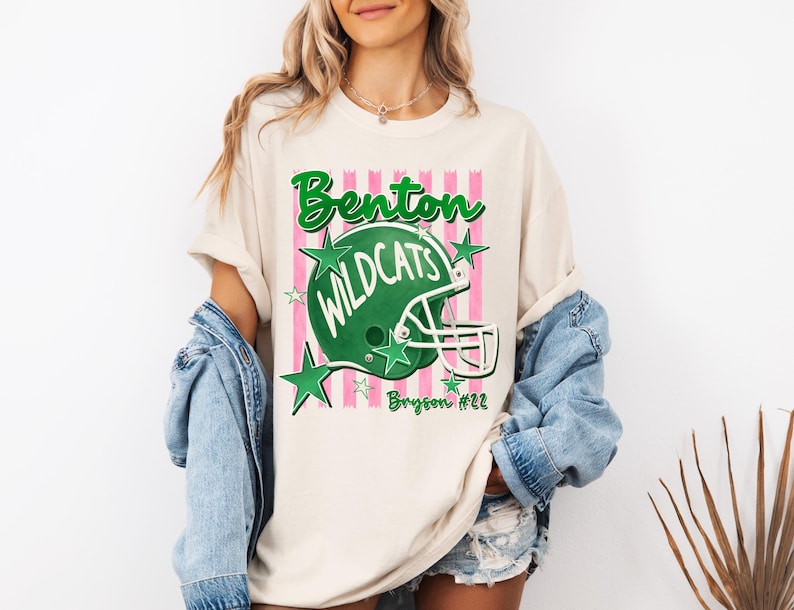K&ouml;nnte beinhalten: Ein cremefarbenes T-Shirt mit einer Grafik eines gr&uuml;nen Football-Helms und dem Text Benton Wildcats. Das Design enth&auml;lt rosa vertikale Streifen, gr&uuml;ne Sterne und den Namen Bryson #22 in gr&uuml;ner Schreibschrift auf der unteren rechten Seite.