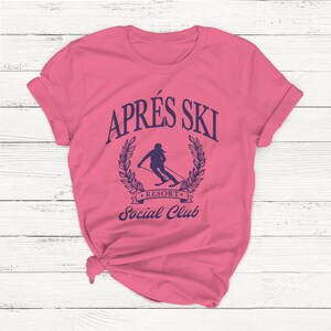 May include: Pink t-shirt with a blue graphic that says "Apr&egrave;s Ski Resort Social Club" and features a silhouette of a skier in a wreath.