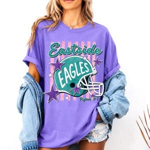 K&ouml;nnte beinhalten: Ein violettes T-Shirt mit einem Grafikdruck eines t&uuml;rkisfarbenen Footballhelms mit dem Text Eastside EAGLES und Peyton #7. Das Design umfasst rosa vertikale Streifen und violette Sterne um den Helm herum, mit einem Rundhalsausschnitt und kurzen &Auml;rmeln.