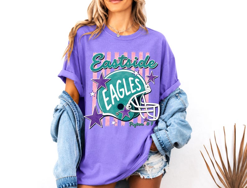 K&ouml;nnte beinhalten: Ein violettes T-Shirt mit einem Grafikdruck eines t&uuml;rkisfarbenen Footballhelms mit dem Text Eastside EAGLES und Peyton #7. Das Design umfasst rosa vertikale Streifen und violette Sterne um den Helm herum, mit einem Rundhalsausschnitt und kurzen &Auml;rmeln.