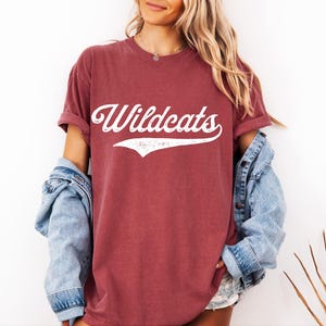 May include: A maroon t-shirt with the word "Wildcats" in white script across the front. The shirt has rolled-up sleeves and is paired with a light blue denim jacket. The overall style is casual and relaxed.