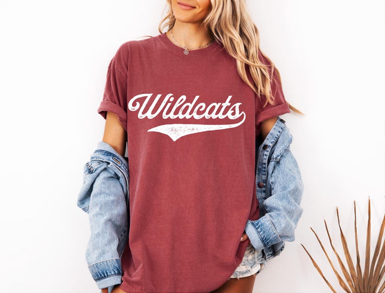 May include: A maroon t-shirt with the word "Wildcats" in white script across the front. The shirt has rolled-up sleeves and is paired with a light blue denim jacket. The overall style is casual and relaxed.