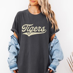 May include: Dark gray t-shirt with the word "Tigers" in a vintage, cream-colored script. The shirt has a relaxed fit and rolled sleeves. A light blue denim jacket is worn over the shirt.