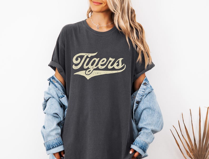May include: Dark gray t-shirt with the word "Tigers" in a vintage, cream-colored script. The shirt has a relaxed fit and rolled sleeves. A light blue denim jacket is worn over the shirt.