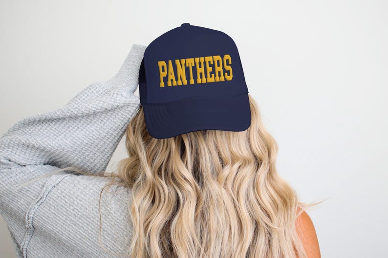 May include: Navy blue baseball cap with the word "PANTHERS" in gold block letters. The cap is worn by someone with long, wavy blonde hair. The background is a plain white wall.