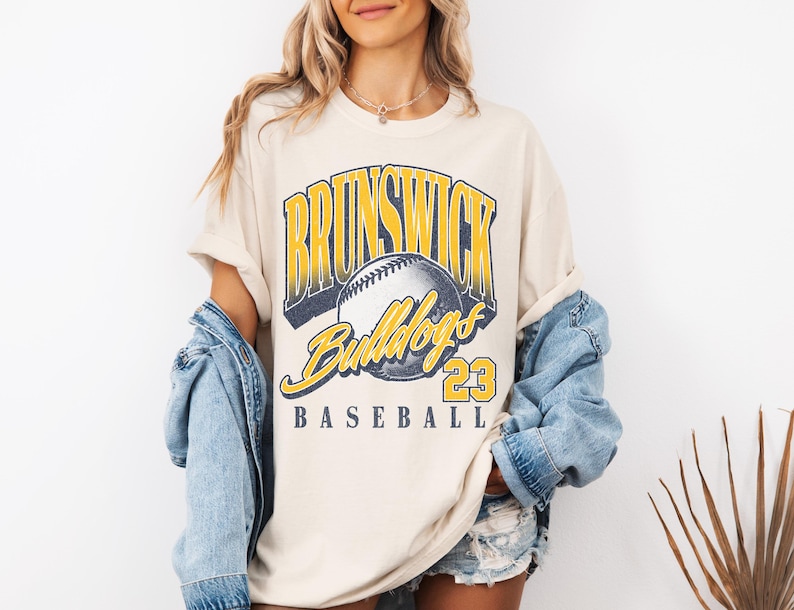 May include: A cream-colored short-sleeve t-shirt featuring a graphic print with the text BRUNSWICK in yellow and blue, a baseball illustration, the word Bulldogs in yellow script, the number 23, and the word BASEBALL in blue text.