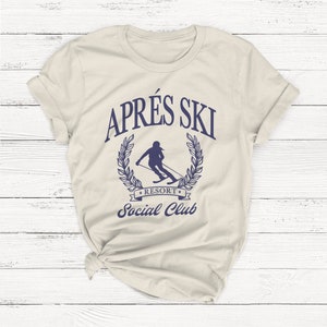 May include: A beige t-shirt with a blue graphic of a skier in a laurel wreath with the text "Apr&egrave;s Ski Resort Social Club".