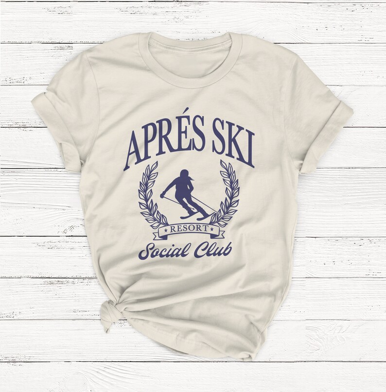 May include: A beige t-shirt with a blue graphic of a skier in a laurel wreath with the text "Apr&egrave;s Ski Resort Social Club".