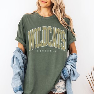 May include: An olive green crewneck t-shirt featuring the word WILDCATS in large, arched, light-colored block lettering with a dark outline, and the word FOOTBALL printed in smaller, simple sans-serif font directly underneath the main text.