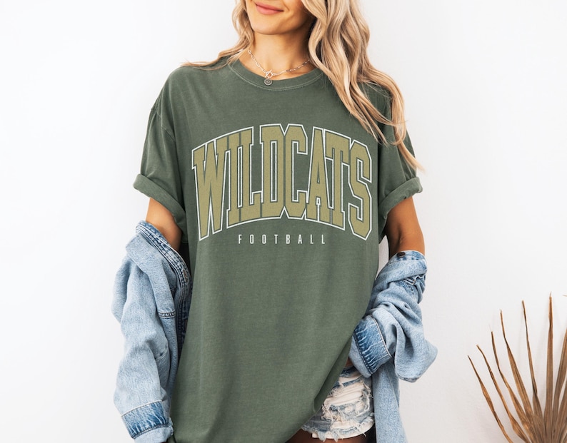 May include: An olive green crewneck t-shirt featuring the word WILDCATS in large, arched, light-colored block lettering with a dark outline, and the word FOOTBALL printed in smaller, simple sans-serif font directly underneath the main text.