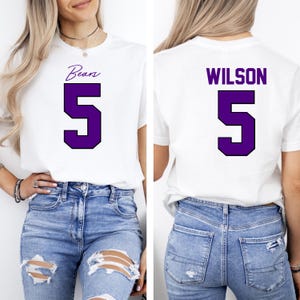 May include: White t-shirt with the word "Bears" and the number "5" in purple on the front. The back of the shirt has the name "WILSON" and the number "5" in purple. The shirt is worn with blue jeans.