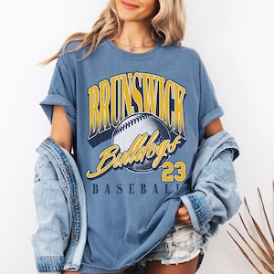 May include: A blue short-sleeve t-shirt featuring a graphic print with the text BRUNSWICK in yellow, Bulldogs in yellow script, and BASEBALL 23 in black. A central illustration of a baseball is included. The shirt has a crew neckline and a relaxed fit.