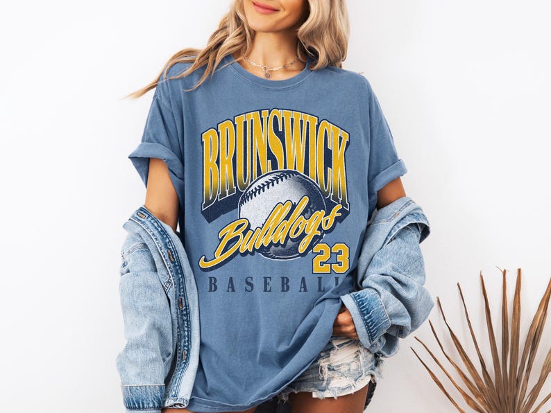 May include: A blue short-sleeve t-shirt featuring a graphic print with the text BRUNSWICK in yellow, Bulldogs in yellow script, and BASEBALL 23 in black. A central illustration of a baseball is included. The shirt has a crew neckline and a relaxed fit.
