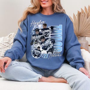 May include: Blue sweatshirt featuring a hockey-themed design. The design includes the name "Haydon" and "Taylor", along with the number "27" and the text "Jay Hawks". The graphic showcases hockey players in action.