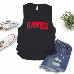May include: Black sleeveless shirt with the word "HAWKS" in red block letters. The shirt is paired with denim shorts and white canvas sneakers. A small green plant is also in the image.