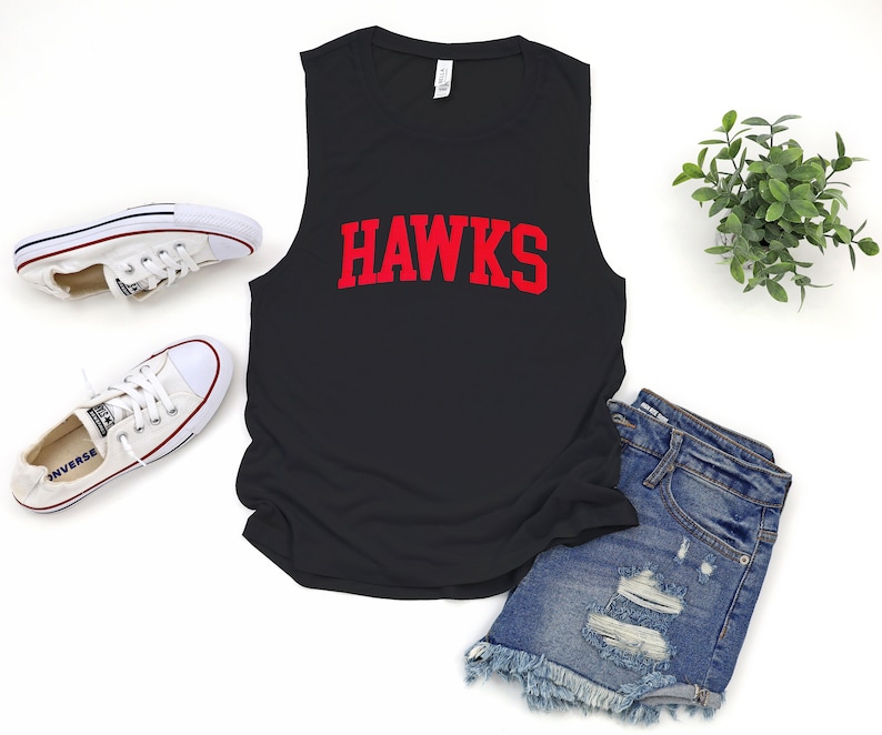 May include: Black sleeveless shirt with the word "HAWKS" in red block letters. The shirt is paired with denim shorts and white canvas sneakers. A small green plant is also in the image.