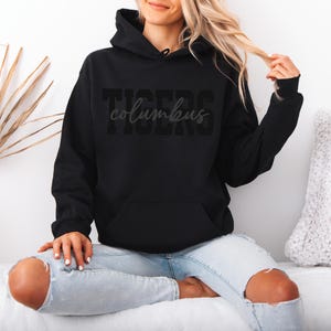 May include: Black hooded sweatshirt with the word "TIGERS" and the word "columbus" printed on the front. The sweatshirt has a front pocket and a drawstring hood. The model is wearing light blue jeans.