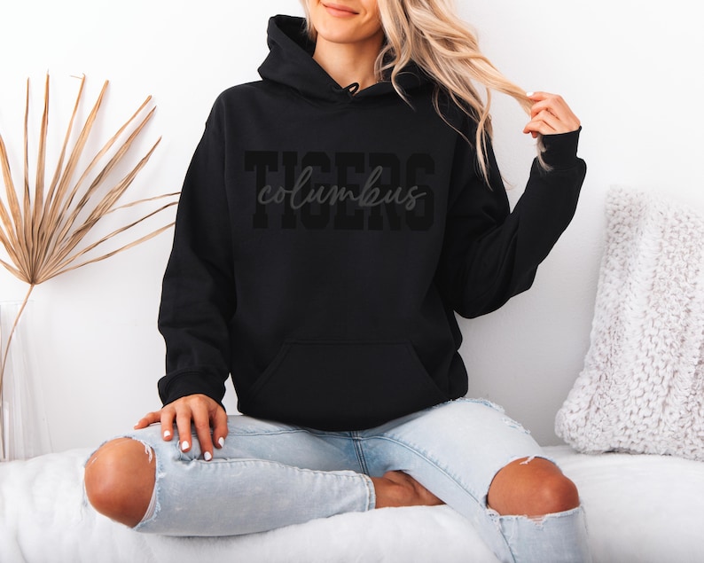 May include: Black hooded sweatshirt with the word "TIGERS" and the word "columbus" printed on the front. The sweatshirt has a front pocket and a drawstring hood. The model is wearing light blue jeans.