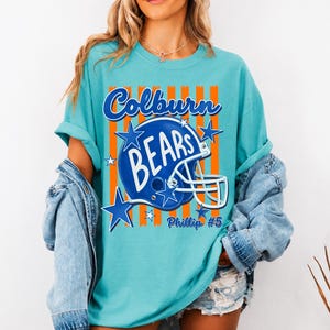 K&ouml;nnte beinhalten: Ein t&uuml;rkisfarbenes T-Shirt mit einem Grafikdruck eines blauen Football-Helms und dem Text Colburn BEARS sowie Phillip #5. Das Design zeigt orangefarbene vertikale Streifen und blaue Sterne auf der Vorderseite, getragen unter einer Jeansjacke.