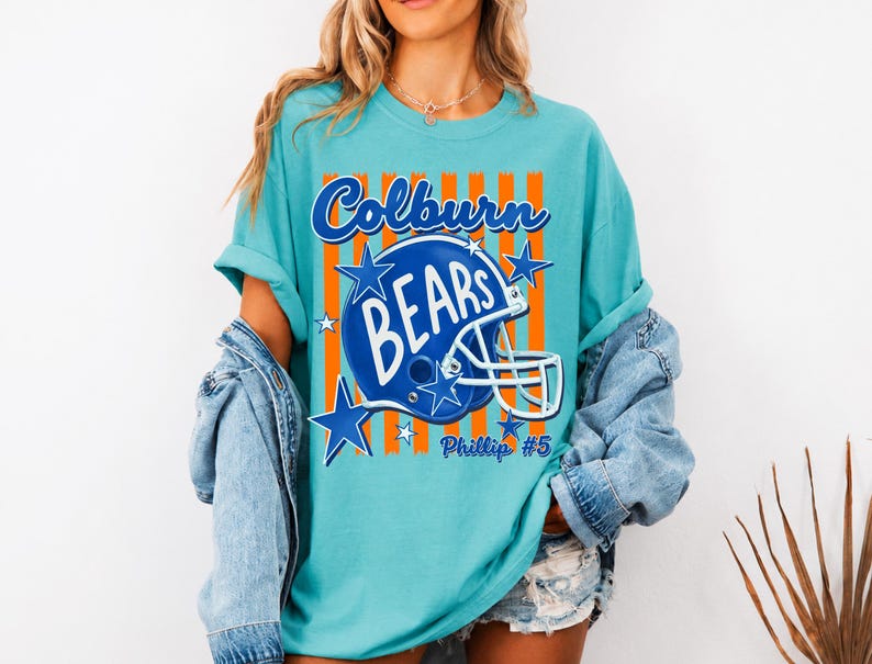 K&ouml;nnte beinhalten: Ein t&uuml;rkisfarbenes T-Shirt mit einem Grafikdruck eines blauen Football-Helms und dem Text Colburn BEARS sowie Phillip #5. Das Design zeigt orangefarbene vertikale Streifen und blaue Sterne auf der Vorderseite, getragen unter einer Jeansjacke.