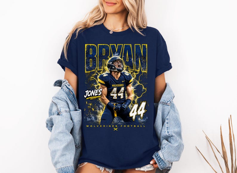 May include: Navy blue t-shirt featuring a football player in a helmet and uniform. The shirt has the name "BRYAN" in yellow, the name "JONES" and the number "44". The text "WOLVERINES FOOTBALL" is also visible.