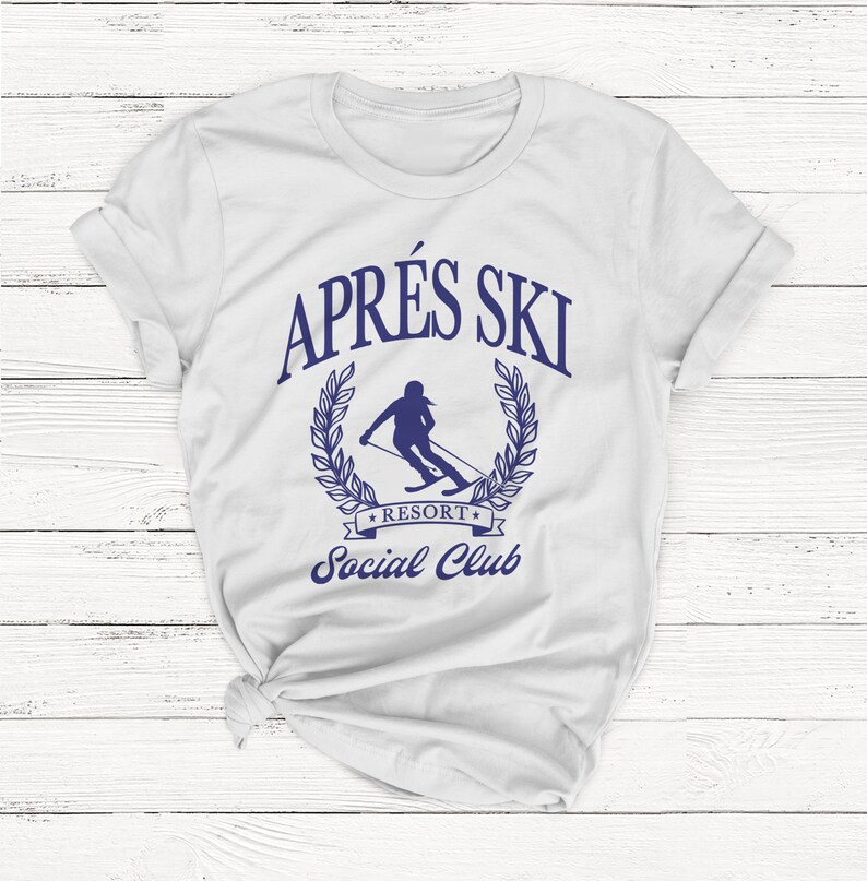 May include: White t-shirt with a blue graphic design that says "Apr&egrave;s Ski Resort Social Club". The graphic features a skier in a wreath.