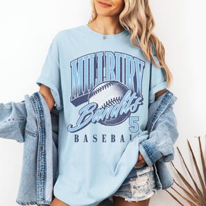 May include: A light blue t-shirt featuring a graphic print of a baseball and the text "MILLBURY BANDITS" in a bold, arched font, with the number "5" and the word "BASEBALL" printed below, paired with a light blue denim jacket and denim shorts.