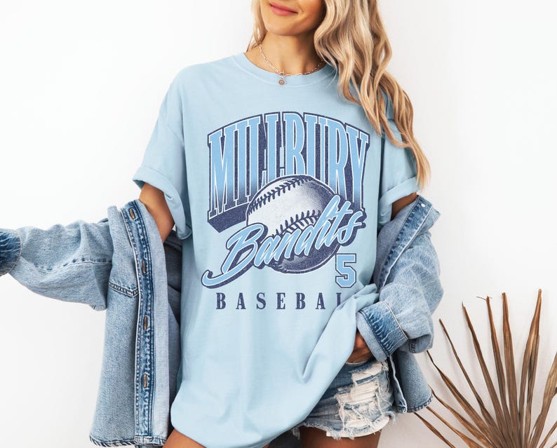 May include: A light blue t-shirt featuring a graphic print of a baseball and the text "MILLBURY BANDITS" in a bold, arched font, with the number "5" and the word "BASEBALL" printed below, paired with a light blue denim jacket and denim shorts.