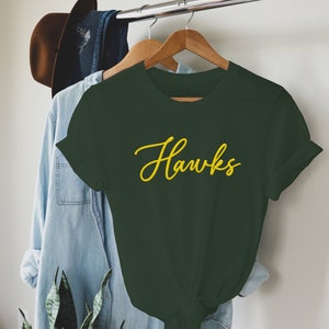 May include: A dark green t-shirt with the word "Hawks" written in yellow cursive lettering.