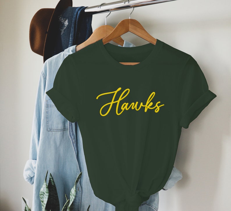 May include: A dark green t-shirt with the word "Hawks" written in yellow cursive lettering.
