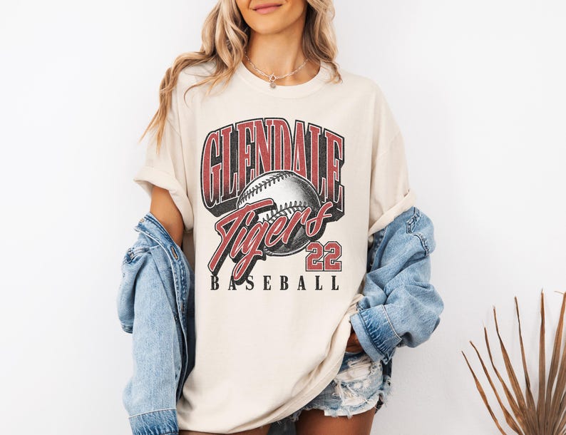 May include: A cream-colored short-sleeve t-shirt featuring a graphic print with the text GLENDALE TIGERS 22 BASEBALL. The design includes a central baseball illustration with red and black lettering, layered over a light blue denim jacket.
