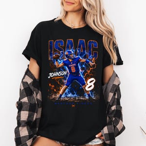 May include: Black t-shirt featuring a football player graphic. The design includes the name "ISAAC" in blue and orange, the name "JOHNSON", the number "8", and the words "MEMPHIS TIGERS". The player is in a blue uniform.