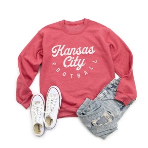 K&ouml;nnte beinhalten: Roter Pullover mit einem wei&szlig;en Aufdruck, der "Kansas City Football" in einer Retro-Schriftart zeigt. Der Pullover wird mit einer Jeans aus abgewetztem Denim und wei&szlig;en Turnschuhen kombiniert.