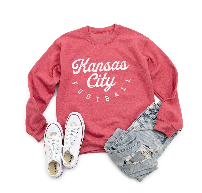 K&ouml;nnte beinhalten: Roter Pullover mit einem wei&szlig;en Aufdruck, der "Kansas City Football" in einer Retro-Schriftart zeigt. Der Pullover wird mit einer Jeans aus abgewetztem Denim und wei&szlig;en Turnschuhen kombiniert.