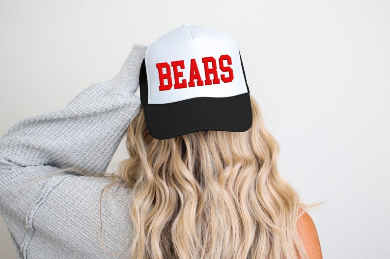 May include: A white and black trucker hat with the word "BEARS" in bold red letters. The hat is worn by a person with long, wavy blonde hair, and a gray sweater. The hat has a mesh back.