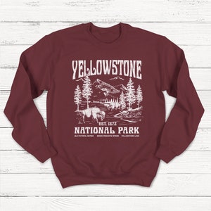 May include: A maroon sweatshirt with a white graphic design of a mountain landscape with trees and a bison. The text "Yellowstone National Park" is printed in white with the words "Est. 1872" below. The text "Old Faithful, Geyser, Grand Prismatic Spring, Yellowstone Lake" is printed in smaller white text below the graphic.