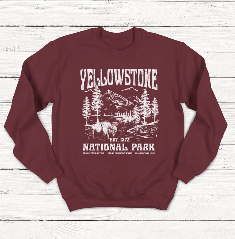 May include: A maroon sweatshirt with a white graphic design of a mountain landscape with trees and a bison. The text "Yellowstone National Park" is printed in white with the words "Est. 1872" below. The text "Old Faithful, Geyser, Grand Prismatic Spring, Yellowstone Lake" is printed in smaller white text below the graphic.