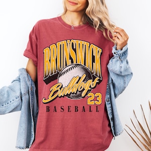 May include: A faded red short-sleeve t-shirt featuring a graphic print with the text BRUNSWICK in yellow, Bulldogs in yellow script, the number 23, and the word BASEBALL in black, centered around a detailed illustration of a baseball.