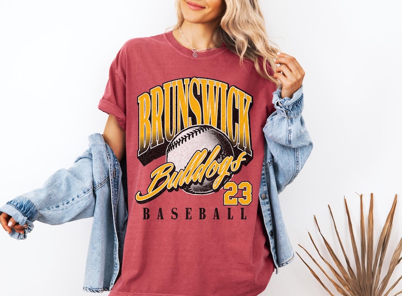 May include: A faded red short-sleeve t-shirt featuring a graphic print with the text BRUNSWICK in yellow, Bulldogs in yellow script, the number 23, and the word BASEBALL in black, centered around a detailed illustration of a baseball.