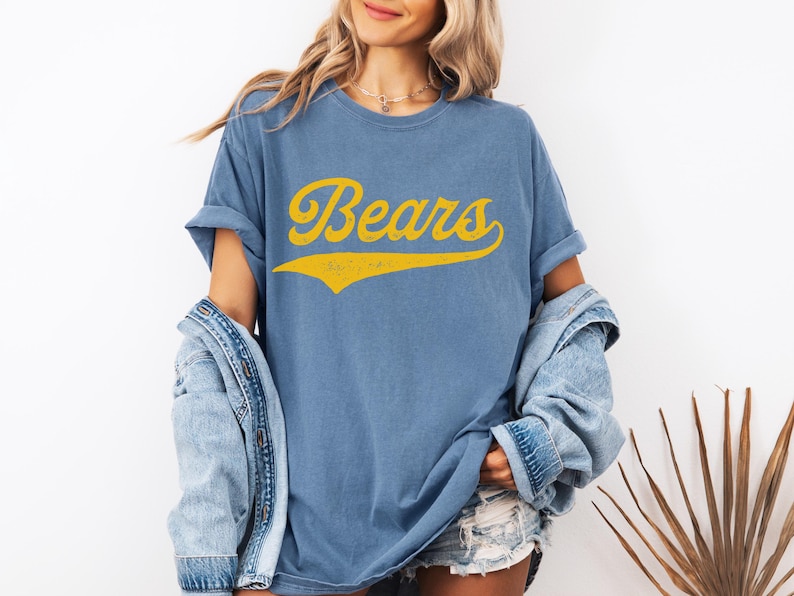 May include: Blue t-shirt with the word "Bears" in yellow script. The shirt has rolled-up sleeves and a vintage look. A denim jacket is draped over the shoulders.