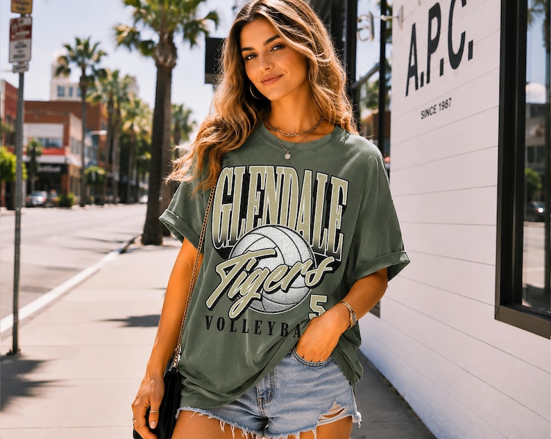 Pu&ograve; includere: Una t-shirt a maniche corte color verde oliva con una stampa grafica di una pallavolo e il testo GLENDALE TIGERS VOLLEYBALL 5 in un carattere audace in stile college. La maglietta ha una vestibilit&agrave; rilassata con maniche risvoltate.