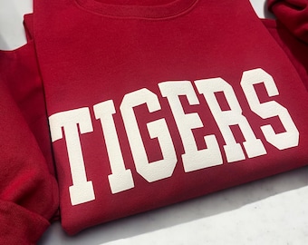 Puede incluir: Sudadera roja con texto blanco que dice "TIGERS" en una fuente en negrita y en bloque.