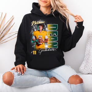 May include: Black hooded sweatshirt featuring a football player in a yellow uniform holding a football. The sweatshirt has the name "Nathan Dean" and the number "11" printed on it, with the word "Grenedoens" below the number.