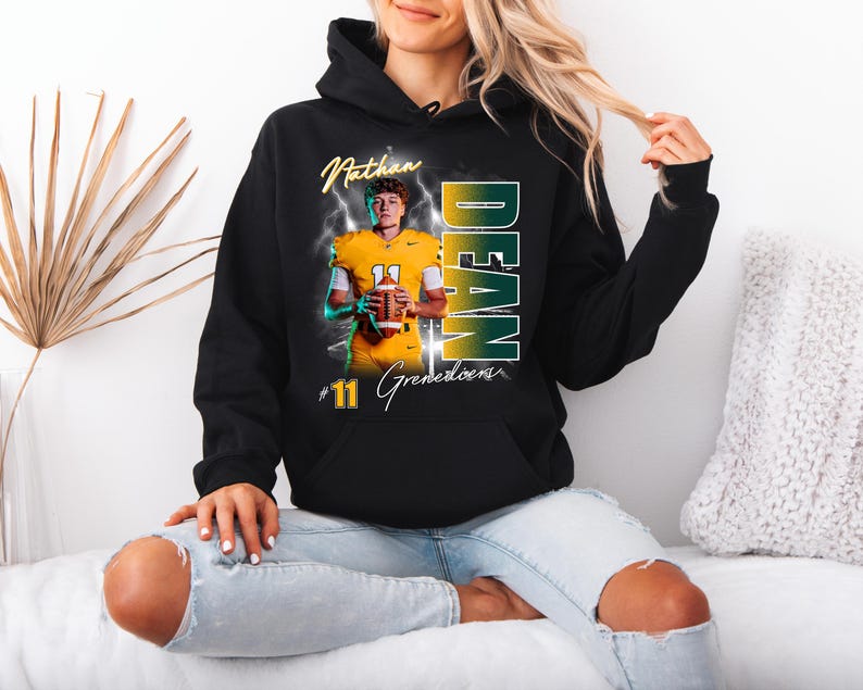 May include: Black hooded sweatshirt featuring a football player in a yellow uniform holding a football. The sweatshirt has the name "Nathan Dean" and the number "11" printed on it, with the word "Grenedoens" below the number.