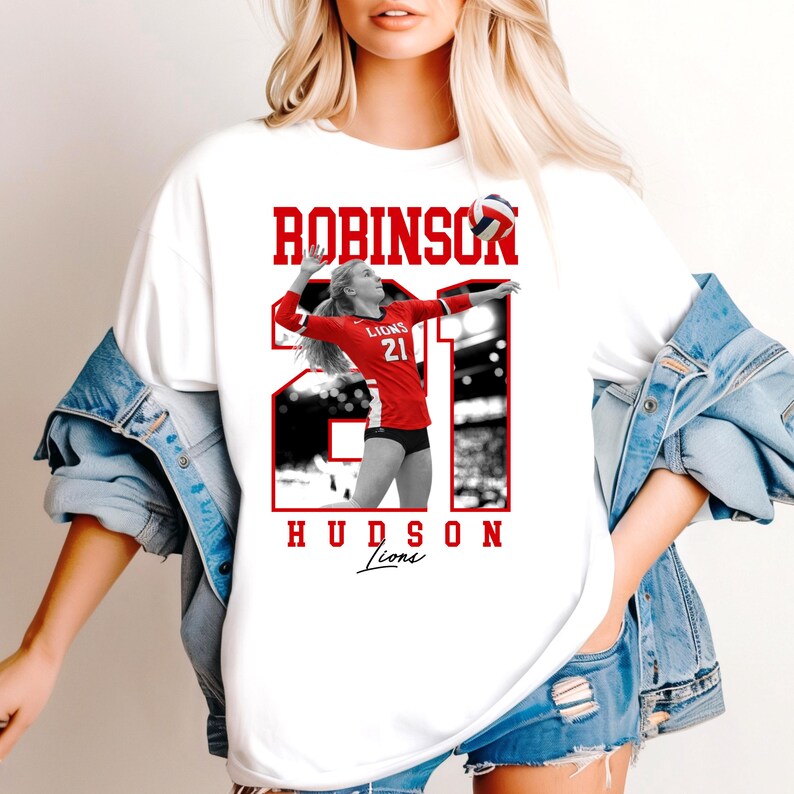 May include: White t-shirt featuring a volleyball player in a red uniform with the number 21. The name "ROBINSON" is printed above the image in red block letters, with "HUDSON Lions" below. The volleyball player is in action, spiking the ball.