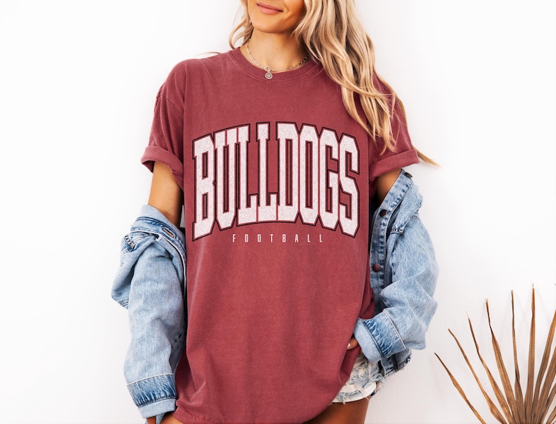 May include: Maroon t-shirt with the word "BULLDOGS" in white block letters with a black outline. Below the word "BULLDOGS" is the word "FOOTBALL" in a smaller font. The shirt has rolled-up sleeves and is paired with a denim jacket.